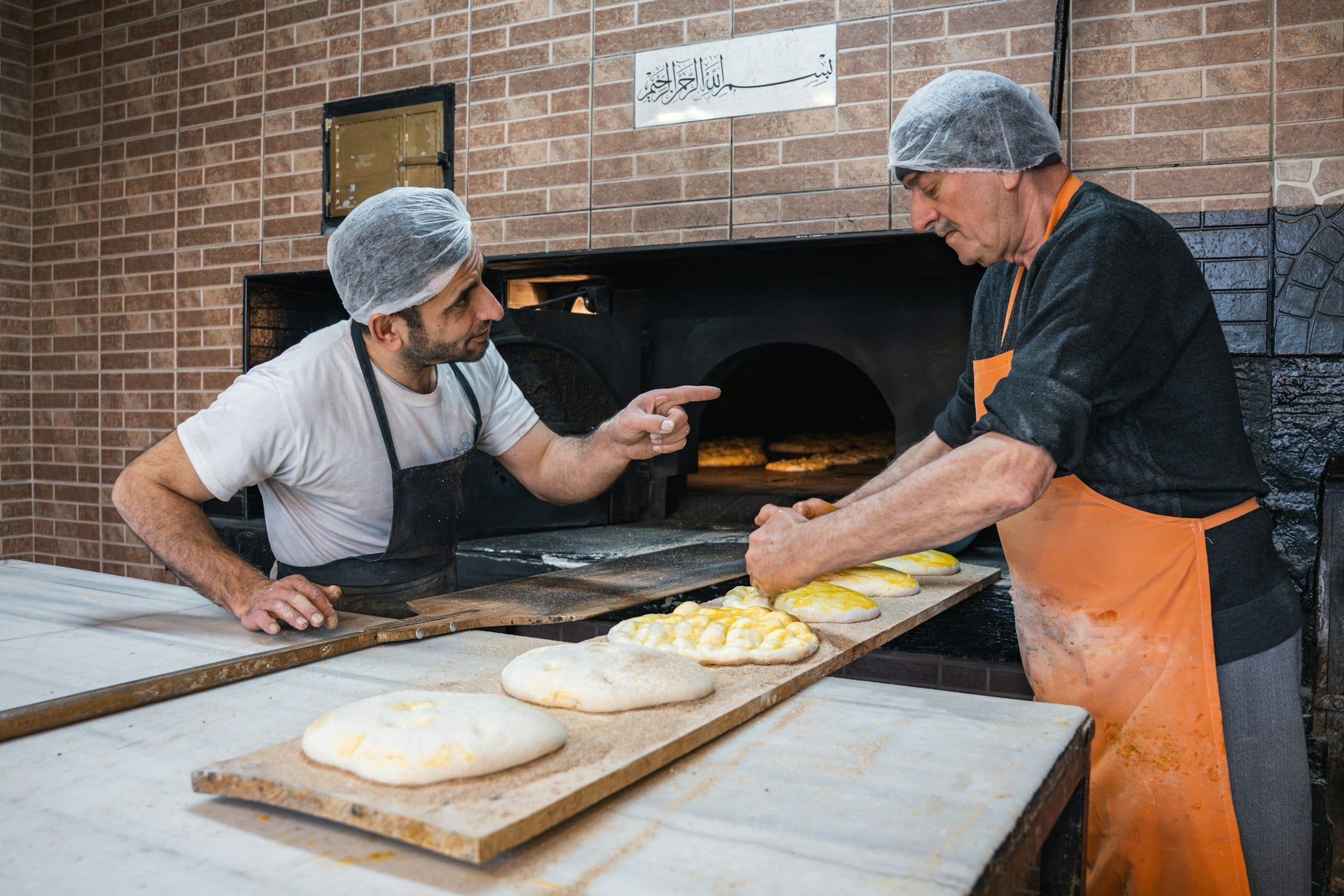 An older baker teaching a younger baker at the oven